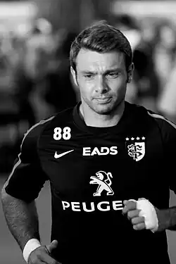 Photographie en noir et blanc, en gros plan, de face, d’un homme portant un maillot foncé, le regard vers le bas, apparemment en train de courir.