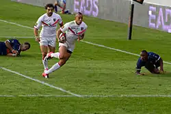 Joueur courant avec le ballon dans une main, suivi par un coéquipier, avec deux adversaires au sol.