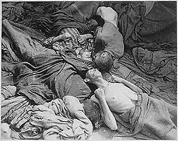 Photo noir et blanc prise dans le camp de Dachau, le 30 avril 1945. D'un fatras de couvertures de différentes nuances de gris, émergent, au centre de la photo, quatre corps décharnés de prisonniers morts de faim. Trois des têtes visibles se rejoignent au centre de la photo.