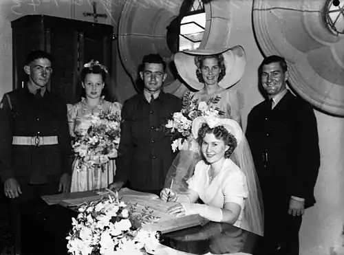 Une jeune femme en robe de mariée signe un registre de mariage en souriant. Derrière, le marié et quatre témoins en tenue militaire.
