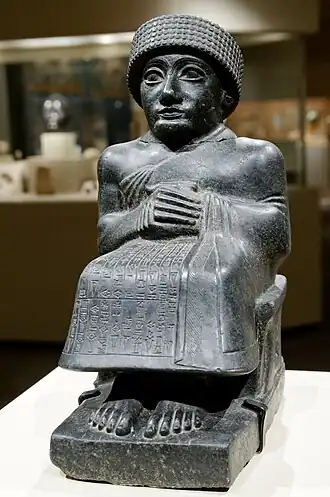 Statuette de pierre noire représentant un homme accroupi, vêtu d'une tunique longue et d'un bonnet.