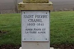 Plaque fixée sur le socle d'une statue (non visible), où est gravé en majuscules "Saint Pierre Chanel 1803-1841 Aimer Marie et la faire aimer".