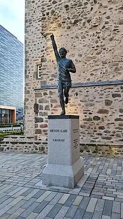 une statue en bronze d'un footballeur, avec un bras levé pointant vers le ciel