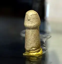 Petite sculpture de pierre beige tubulaire en forme de têtard. La partie haute évoque un visage grossièrement gravé.