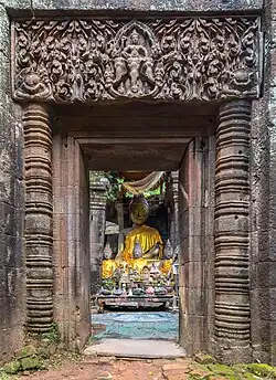Porte en pierre avec des colonnes et des reliefs bouddhistes, menant à une pièce avec une statue de Bouddha assis vêtu d'une tunique orange, une table avec des pierres et des reliques bouddhistes, dans le complexe du temple hindou khmer de Wat Phou. Septembre 2019.