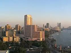 High-rises, including a multi-story white building in the foreground, dominate a sunset view of Cairo alongside the Nile, which is shown flowing under a bridge carrying a busy street