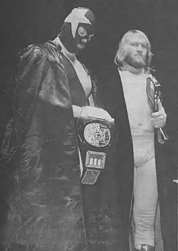 Une photographie en noir et blanc de deux catcheurs, un d'entre eux porte un masque avec une étoile et l'autre a des cheveux blond mi-long et une barbe. Ils ont tous les deux une ceinture de champion de catch.