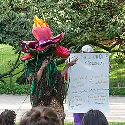 Photographie en plein air. Une artiste à gauche porte un costume de plante, avec un chapeau en forme de fleur rose ouverte et une tenue verte et marron en crochet. À sa droite, une personne tient une grande pancarte "né de l'ordre colonial. Contrôle du vivant = maximiser l'exploitation. sexe, genre, corps, environnement. Hijras : Inde, Pakistan. Code Pénal Colonial § 145 8 148 Ouganda."