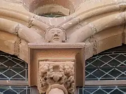 Photo couleur montrant un chapiteau de fenêtre sculpté de feuilles de vigne et surmonté d'une tête humaine et d'un oculus. Les ouvertures sont dotées de panneaux de vitraux de verre en losange.