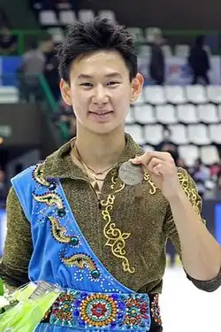 Un homme asiatique souriant en costume brun, vert et bleu, avec une médaille.