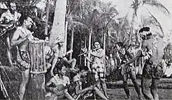 Photo en noir et blanc d'un groupe de personnes d'origine insulaire qui participe à une performance culturelle, en vêtements traditionnels légers. Ils jouent des instruments comme des tambours et des flûtes, et certains dansent. Le décor naturel, avec des arbres tropicaux, crée une ambiance joyeuse, évoquant un rituel ou un spectacle.