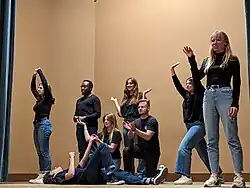 Plusieurs personnes se tiennent debout en levant les bras. D'autres personnes se tiennent devant elles, à genoux ou allongé au sol, en faisant des gestes également.