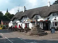 Photo d'une grande croix en pierre sur un piédestal de quelques marches devant une grande maison aux murs blancs