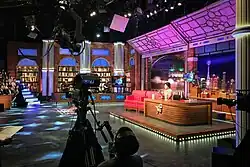 Photographie couleurs d'un immense plateau TV très coloré, une caméra en gros plan. une femme brune est assise derrière un grand bureau de bois