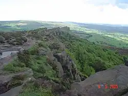 au premier plan, le sommet d'une falaise composée de vastes blocs gris entrecoupés de buissons, dominant un paysage de bocage qui s'étend en arrière plan