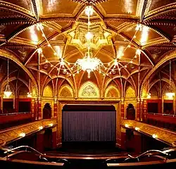 Photographe d'une grande salle comportant une scène avec un rideau foncé, des boiseries et un chandelier au plafond.