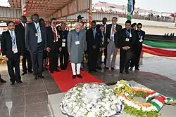 Photographie en couleurs de plusieurs hommes, pour la plupart en tenue noire, se recueillant devant deux grandes couronnes de fleurs.