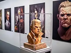 Exposition dans une galerie d'un buste d'homme aux traits burinés et agressifs et de plusieurs portraits de personnages élégants au regard froid.