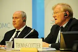 Photographie de deux hommes, celui à droite au premier plan, portant des lunettes, parle dans un micro, celui à gauche en arrière plan, réfléchit.