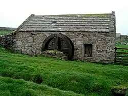 La photo montre un bâtiment en pierre d'un âge certain. Au milieu du bâtiment se trouvent les restes d'une roue en bois.