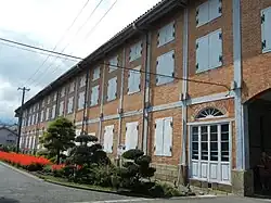 Photo couleur de la façade d'un entrepôt à deux étages, construction en briques rouges aux volets blancs fermés. Devant le bâtiment : un arrangement floral fait d'arbustes au feuillage vert et de massifs de fleurs rouges. Un ciel nuageux en arrière-plan.