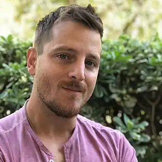 Photo en buste d'un homme châtain portant un haut mauve et souriant.