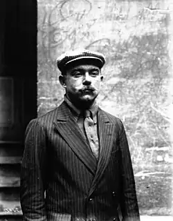 Portrait d'un homme vêtu d'un costume et d'une casquette, se tenant debout.