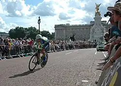 Photographie couleur d'un cycliste sur son vélo, en tenue de cycliste bleue et verte, salué par la foule dans une rue de Londres, sous un ciel bleu nuageux.