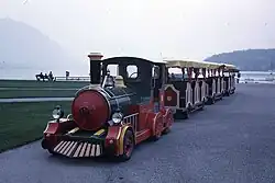 Photographie en couleurs d’un petit train sur pneus multicolore avec trois wagons rouge et blancs sur une route au bord de l’herbe. En arrière-plan, on distingue un lac et des montagnes sous un ciel laiteux.