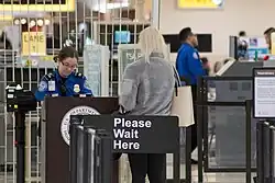 Photo d'une femme de dos qui se tient devant un comptoir de contrôle de sécurité dans un aéroport. Une agente en uniforme bleu est assise derrière le comptoir, vérifiant ses documents. Un panneau sur le comptoir indique "Please Wait Here". En arrière-plan, d'autres agents et passagers sont visibles près des files d'attente et des équipements de sécurité.