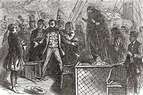 Gravure, Greenwood écartant les bras devant une table sur laquelle se tient une femme voilée.