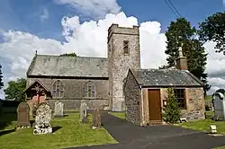 Photo d'une église vu de côté comprenant une petite tour attachée à un bâtiment moins haut. Devant l'église, sur la gauche, se trouvent des pierres tombales alors que sur la droite se trouve une petite annexe avec une porte marron.