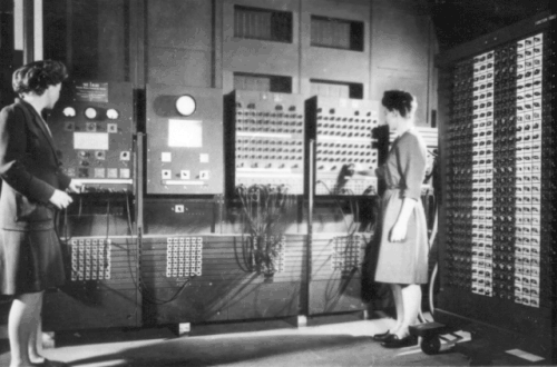 Au fond de la pièce, une grosse machine, l’ordinateur ENIAC. La machine est composée de 7 panneaux, recouverts de boutons, d’indicateurs lumineux et de câbles. On peut voir un amas de câbles branchés sur l’ordinateur. Devant la machine se tiennent debout deux femmes, Betty Jenning à gauche et Frances Bilas à droite. Frances Bilas règle un bouton rotatif, tandis que Betty Jenning semble bouger des câbles.