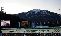 Une foule multicolore regarde passer un bobsleigh noir et blanc sur une piste de bobsleigh marquée des anneaux olympique et de grandes lettres «&nbsp;vancouver 2010&nbsp;» ; derrière, se trouve un écran géant vidéo retransmettant la scène, un autre écran détaillant des résultats sportifs, une rangée de bannières olympiques et de plusieurs pays ; encore plus loin, un paysage de petite montagne partiellement enneigée, et un petit dirigeable d'observation dans le ciel