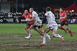 joueur de profil, portant le ballon, deux adversaires courant dans le même sens que lui. Un coéquipier, de dos, vient à sa rencontre.