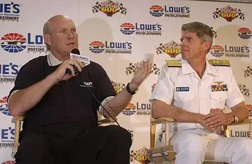 Un homme chauve habillé de noir tient un micro et discute sous les yeux d'un militaire paré de blanc.