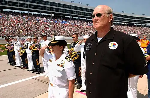 Un homme imposant se tient droit, debout, devant des militaires au garde à vous et une foule massée en tribune.