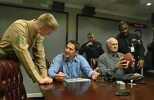 Plusieurs hommes autour d'une table discutent. L'un d'eux tient un ballon de football américain dans ses mains.