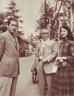 Photographie en noir et blanc de trois individus japonais, qui regardent l'objectif avec le sourire, deux hommes et une femme. Les deux hommes sont en costumes. La scène a lieu dans une rue, le jour.