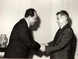 Ieng Sary à gauche serre les mains de Nicolae Ceaușescu à droite; à l’extrême gauche un vase trône sur un guéridon