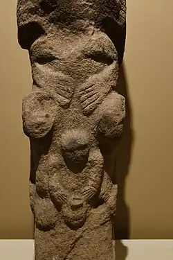 Pierre oblongue beige sculptée, posée sur une de ses petites faces carrées. Elle est posée sur un socle blanc face à un mur beige plus foncé. Le tout est éclairé par un projecteur électrique.