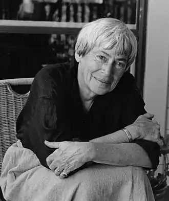 Photo portrait en noir et blanc d'Ursula Le Guin qui est assise et croise les bras devant une étagère de livres.