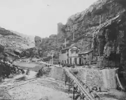Photographie en noir et blanc d'un bâtiment surplombant un cours d'eau et surmonté de plusieurs conduites forcés.