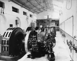 Photographie en noir et blanc de l'intérieur d'un bâtiment abritant quatre alternateurs de forme cylindrique.