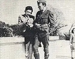 Deux personnages sont sur la photo. La femme est assis sur un petit muret, l'homme est debout à côté d'elle, devant le muret. Les deux personnes sont en tenues militaires. La photo est en noir et blanc.