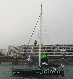 Voilier noir, de profil, progressant au moteur dans un chenal.