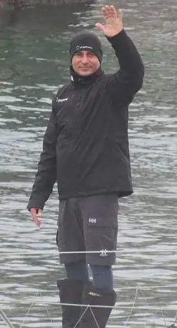 debout sur le pont de son bateau, le navigateur, habillé de noir, salue la foule.