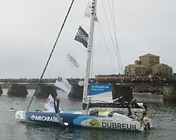 Foiler bleu et blanc progressant au moteur le long d'une estacade où la foule est massée. À l'avant, le navigateur salue la foule.