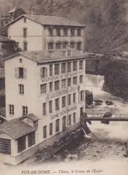 Carte postale ancienne en bistre et blanc montrant le centre de la vallée avec la Durolle juste en avant de la cascade et sur la gauche les bâtiments de l'usine d'Entraygues s'élevant en un parallélépipède de 4 étages à 7 fenêtres rectangulaires par niveau. On lit très clairement sur les ceintures de maçonnerie entre les étages de haut en bas : Manufacture de Coutellerie Fine, Sauzede Père et Fils, Usine d'Entraygues
