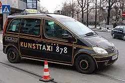 Photo d'une voiture noire avec des lignes jaunes, sur lequel est posée une plaque de taxi, traversant une ligne de tramway.